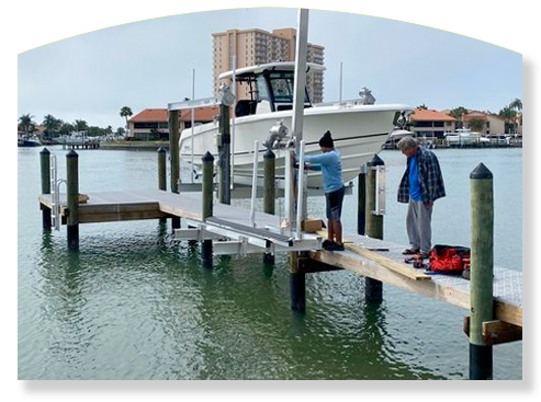 Two people installing a boat lift on a dock. Boat is being lifted out of the water. Buildings in the background.