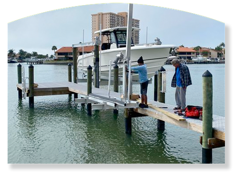 Boat being lifted by a lift at a dock. Two people work on the lift. Water, buildings, and sky in the background.