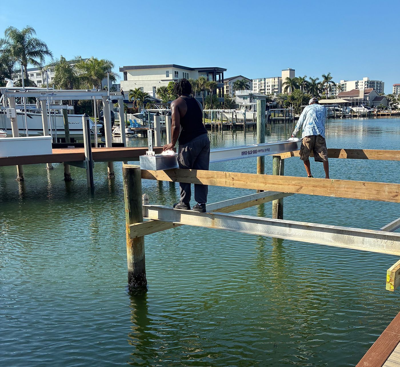 Two people on a pier working on dock construction. Sunny day, water, buildings in background.