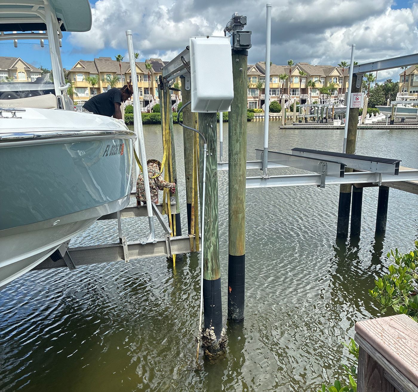 Boat on a lift at a dock; two people working on the boat near waterfront condos.