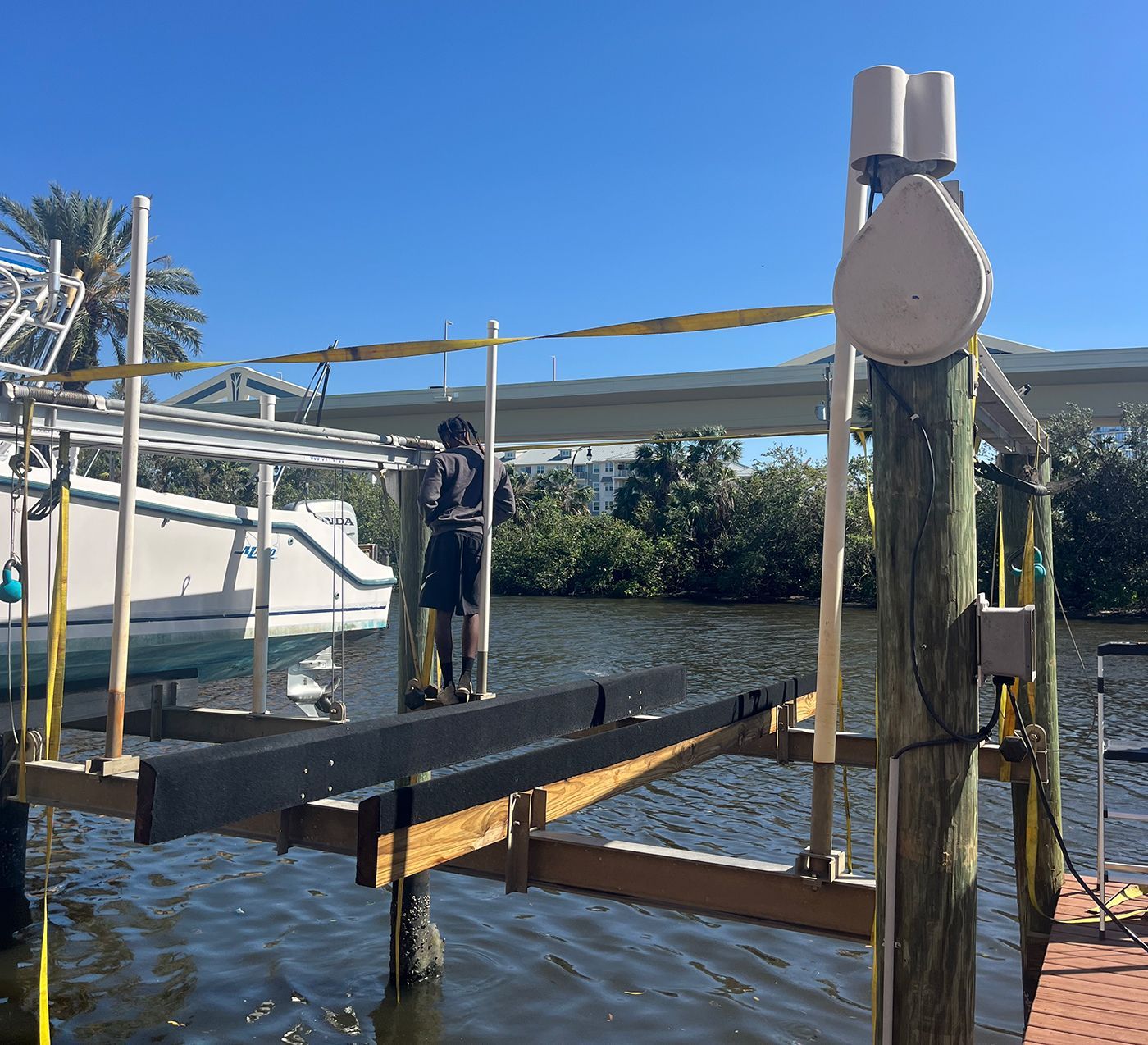 Person on a boat lift, adjusting supports. Wooden dock, boat, and water in the background. Sunny day.