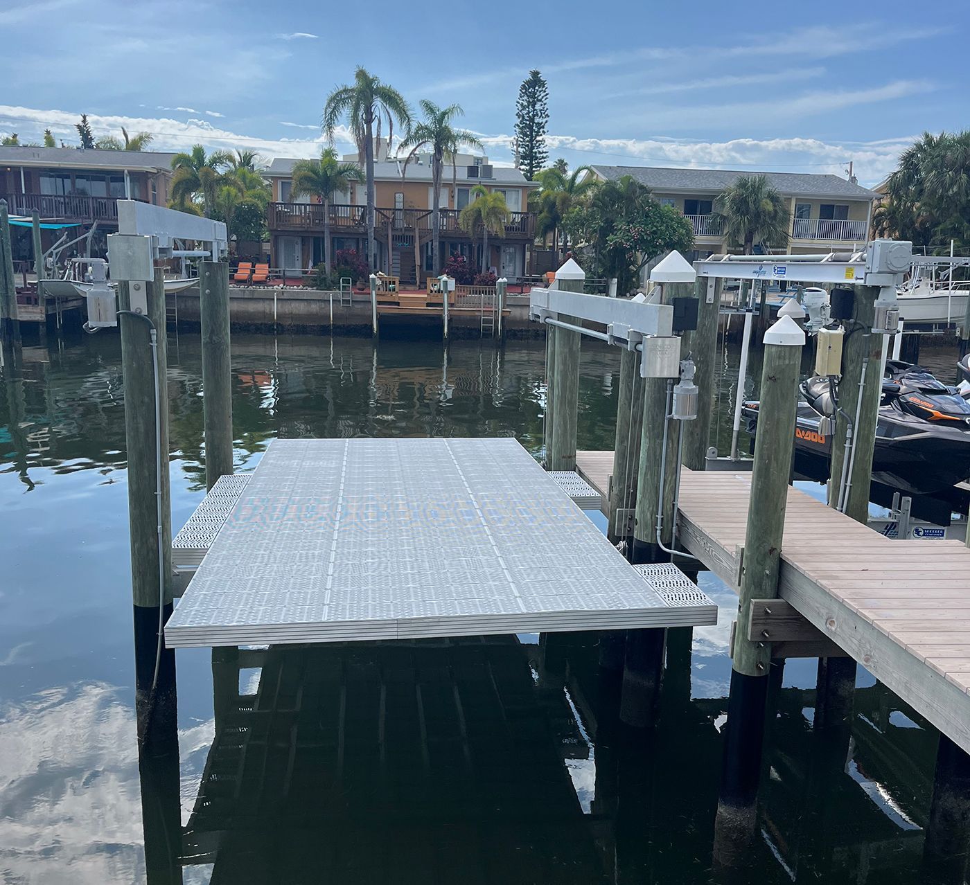 Dock with a silver ramp leading to a pier on a canal. Residences and palm trees are in the background.