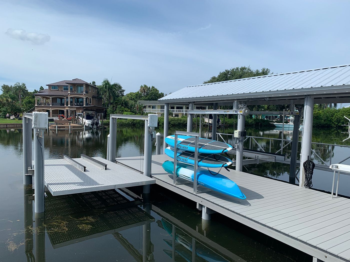 Dock with kayaks and boat lift under a metal roof, with a large house in the background.