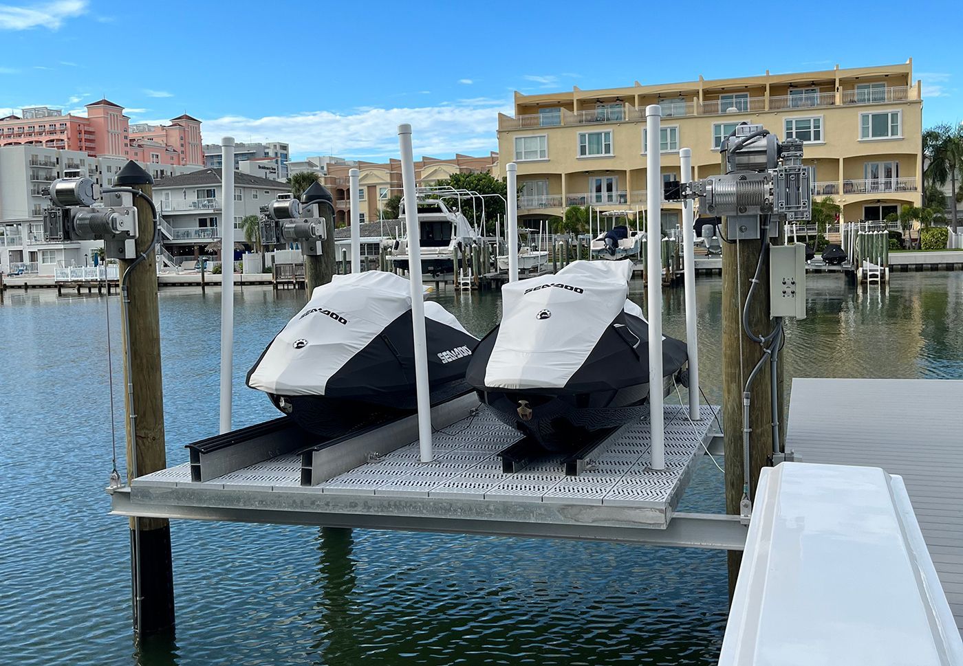 Two boats covered with black tarps on a boat lift at a dock. Buildings and water in the background.