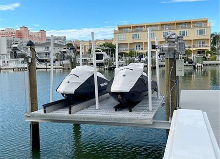 Two covered jet skis on a boat lift at a marina, with waterfront condos in the background.