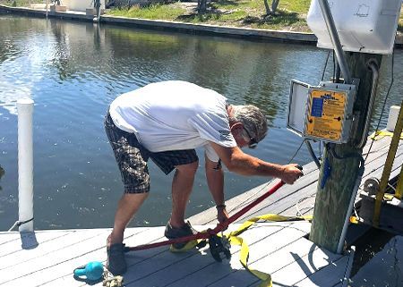 Man using a red tool on a wooden dock near a canal. The man wears a white shirt and shorts.