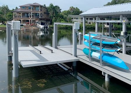 Dock with paddleboards, lift, and boat; a large house is in the background on a waterway under a partly cloudy sky.