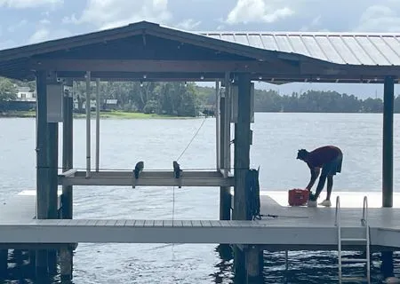 A person in a dark shirt and shorts stands on a wooden dock under a boat lift cover, leaning over to reach into a red bin.
