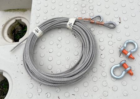 Coiled steel cable with attached eyelets and two shackles, resting on a white, textured surface.
