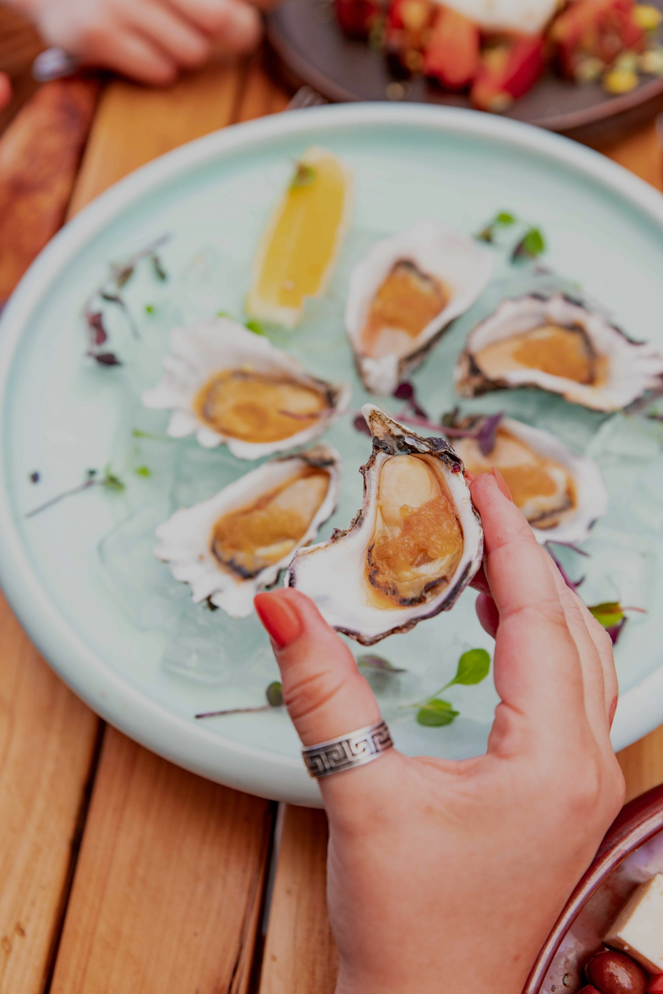 Hand holding a fresh oyster over a plate of oysters with lemon wedges and sauce on a wooden table