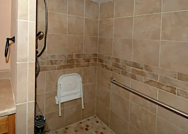 A walk in shower with a folding chair and a stainless steel railing.