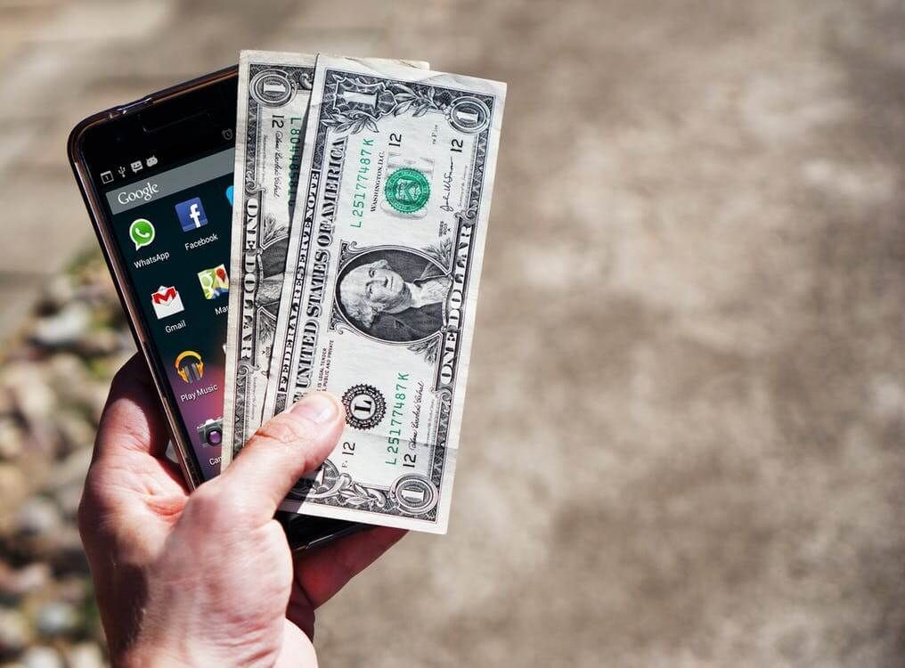 A person is holding a cell phone and a dollar bill.