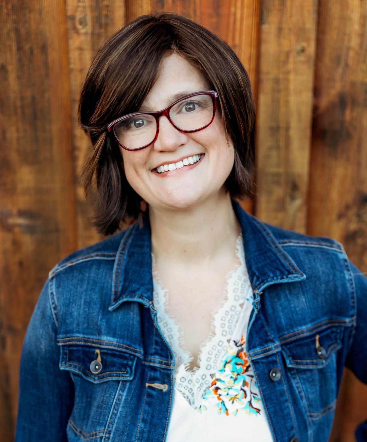 A woman wearing glasses and a denim jacket is smiling in front of a wooden wall.