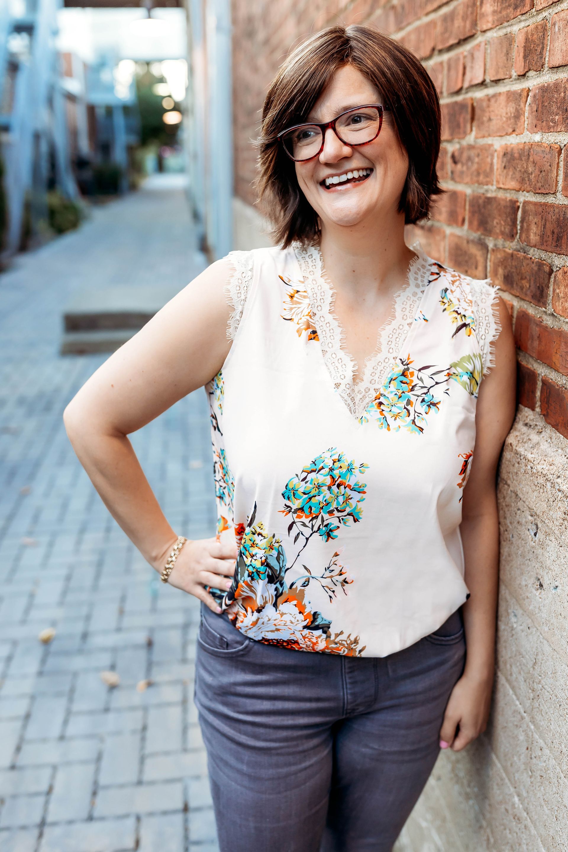 A woman wearing glasses is leaning against a brick wall and smiling.