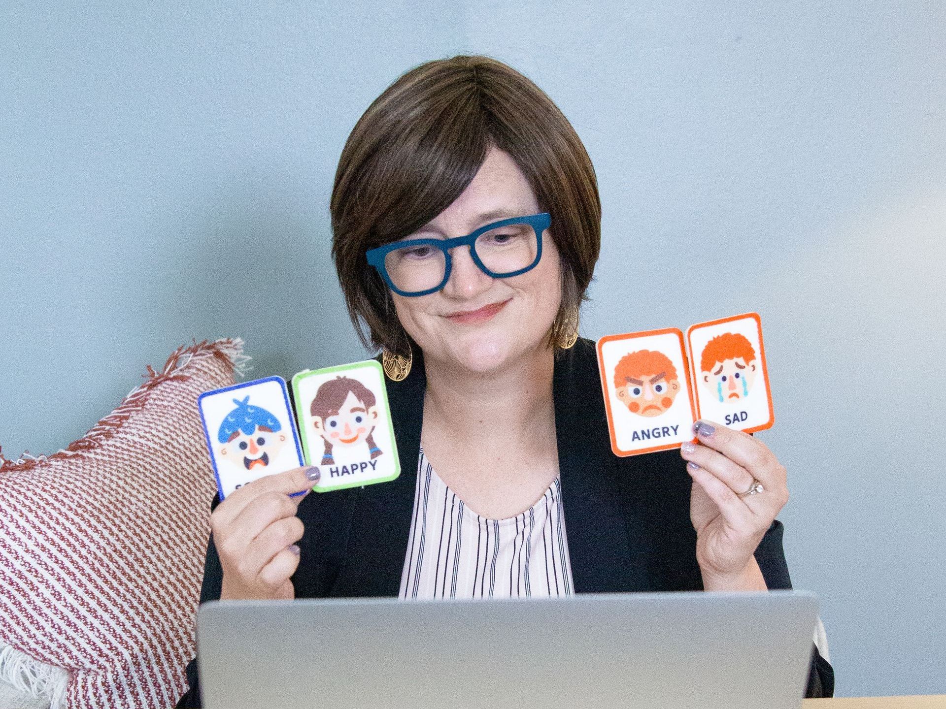 A woman wearing glasses is sitting in front of a laptop holding cards with cartoon faces on them.