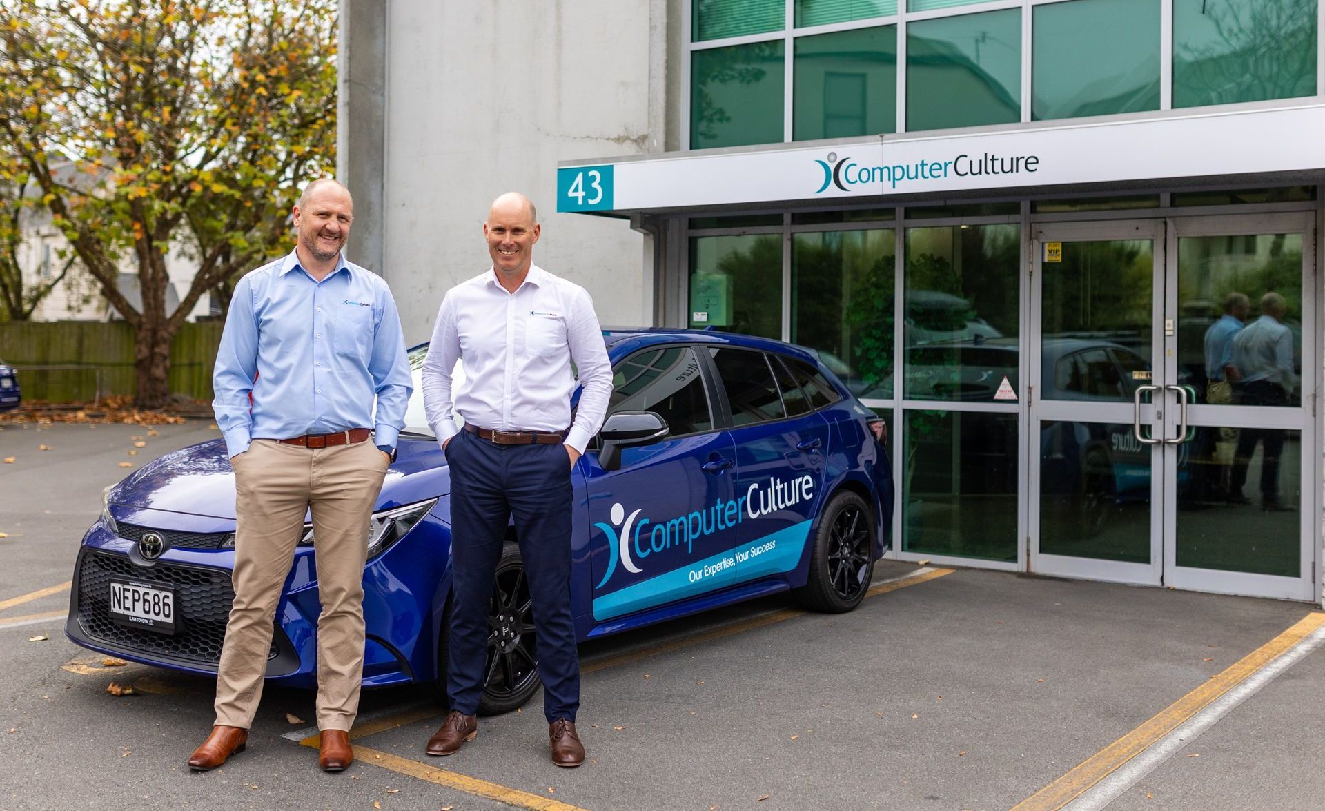 Craig Harrington and Kevin Riddle, Computer Culture founders and directors standing in front of a blue Computer Culture branded car. Outside of the Computer Culture building.