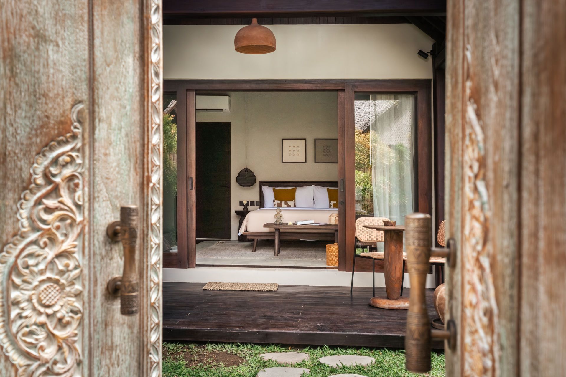 Open wooden doorway framing a cozy bedroom with a bed and chairs on a porch-like terrace.