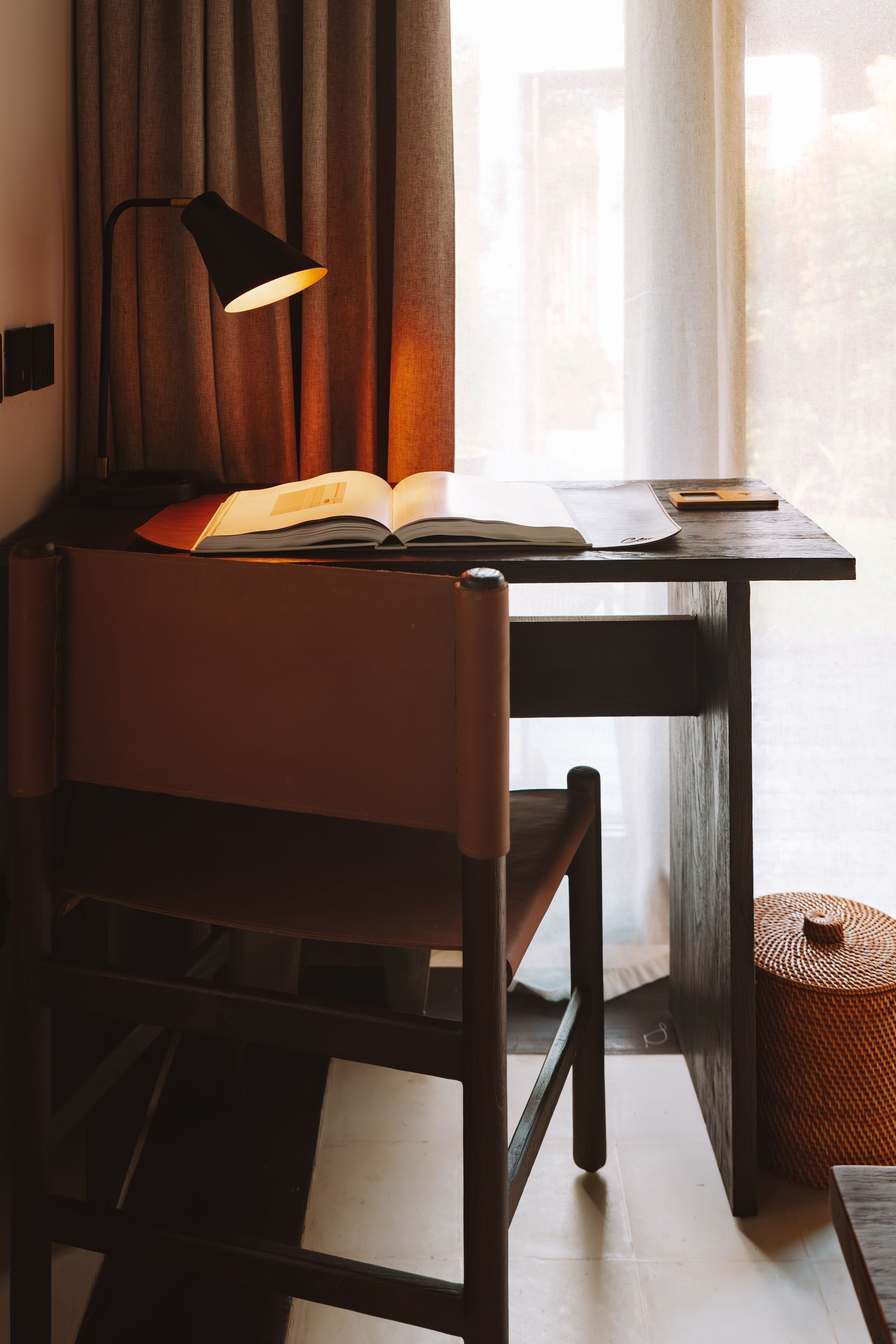 Cozy desk by a window with an open book, lamp, chair, and wicker basket in warm light
