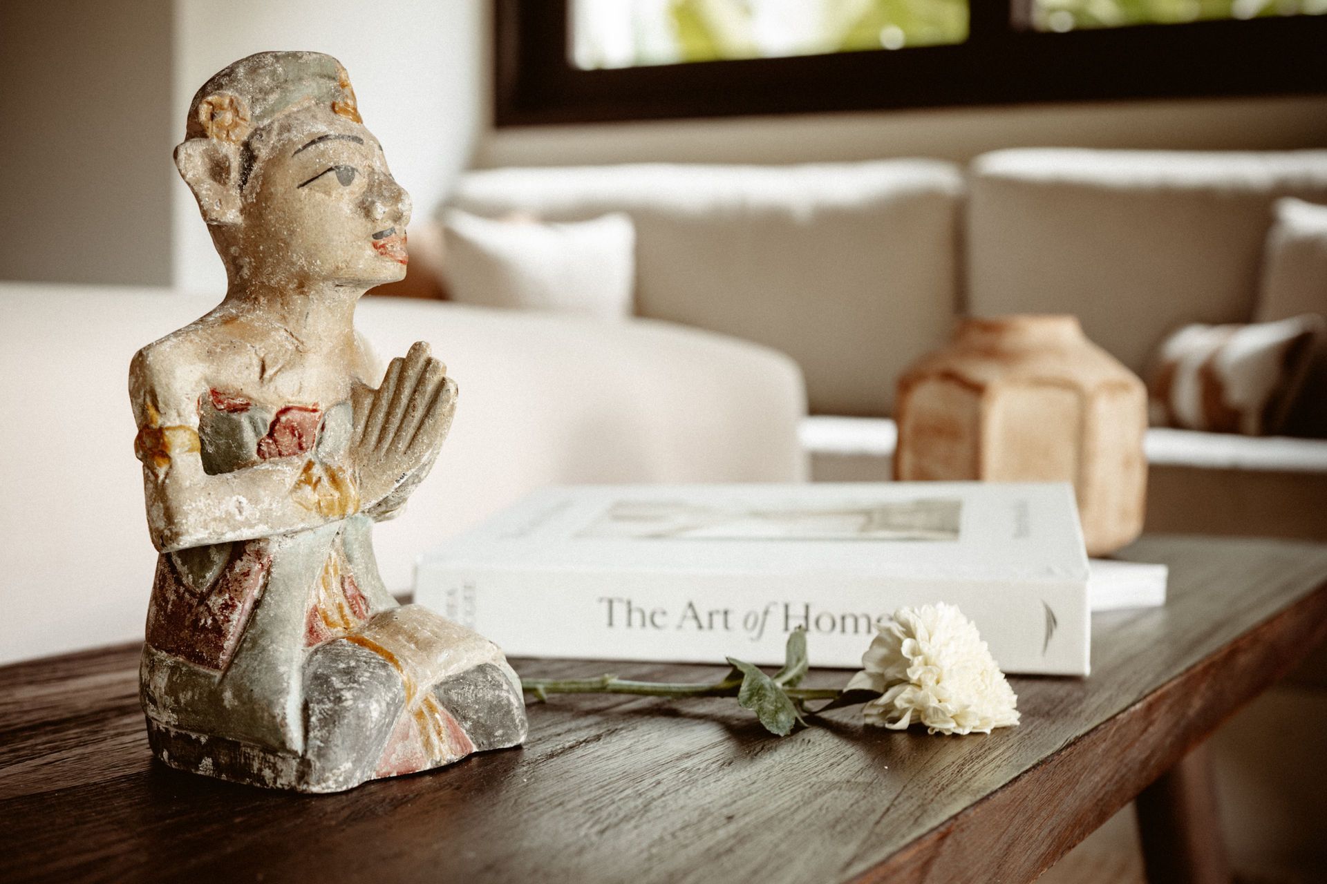 A small stone statue and white flower on a wooden coffee table beside a book in a bright living room