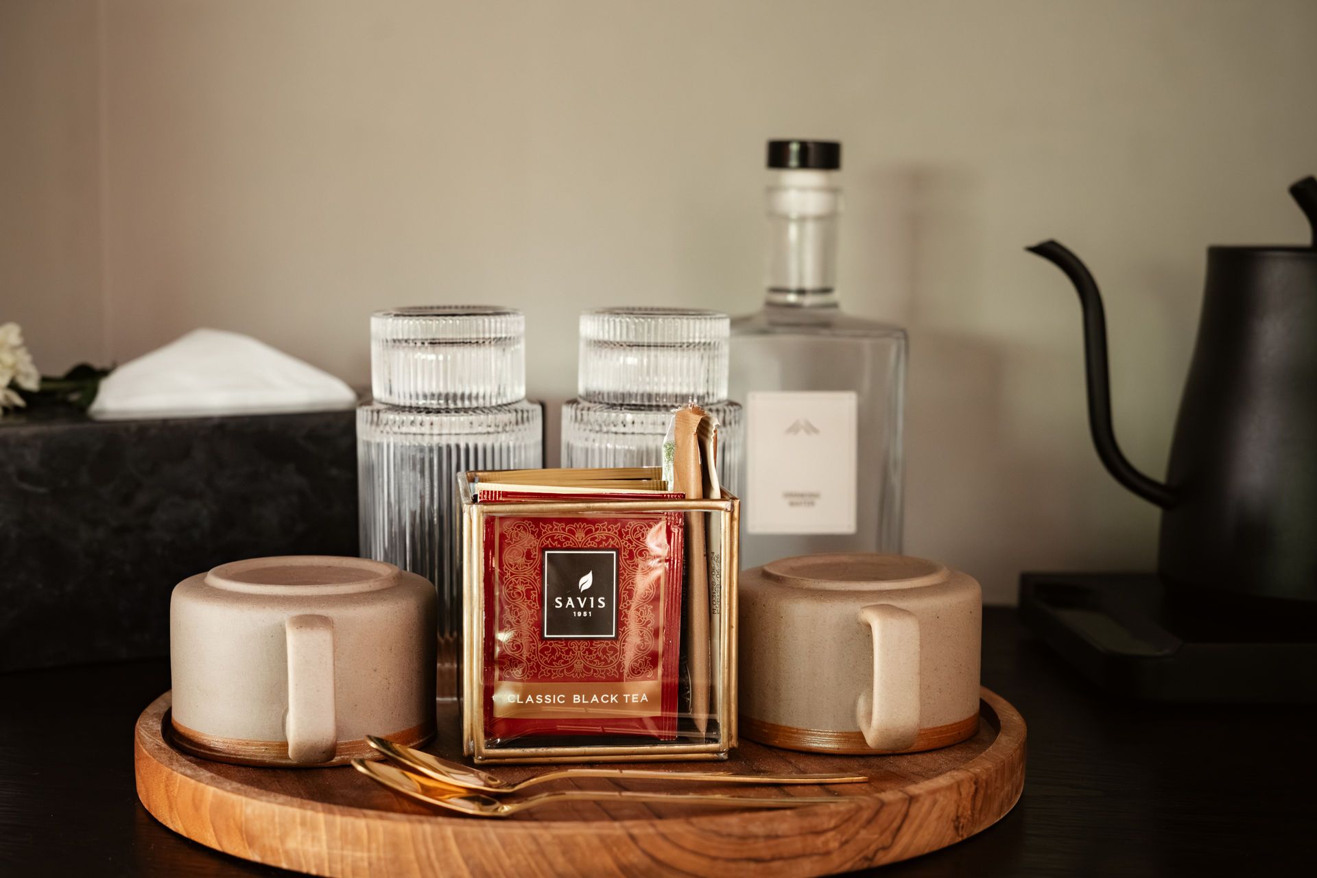 Candles, a red-labeled bottle, and glassware arranged on a wooden tray, with a black kettle in the background