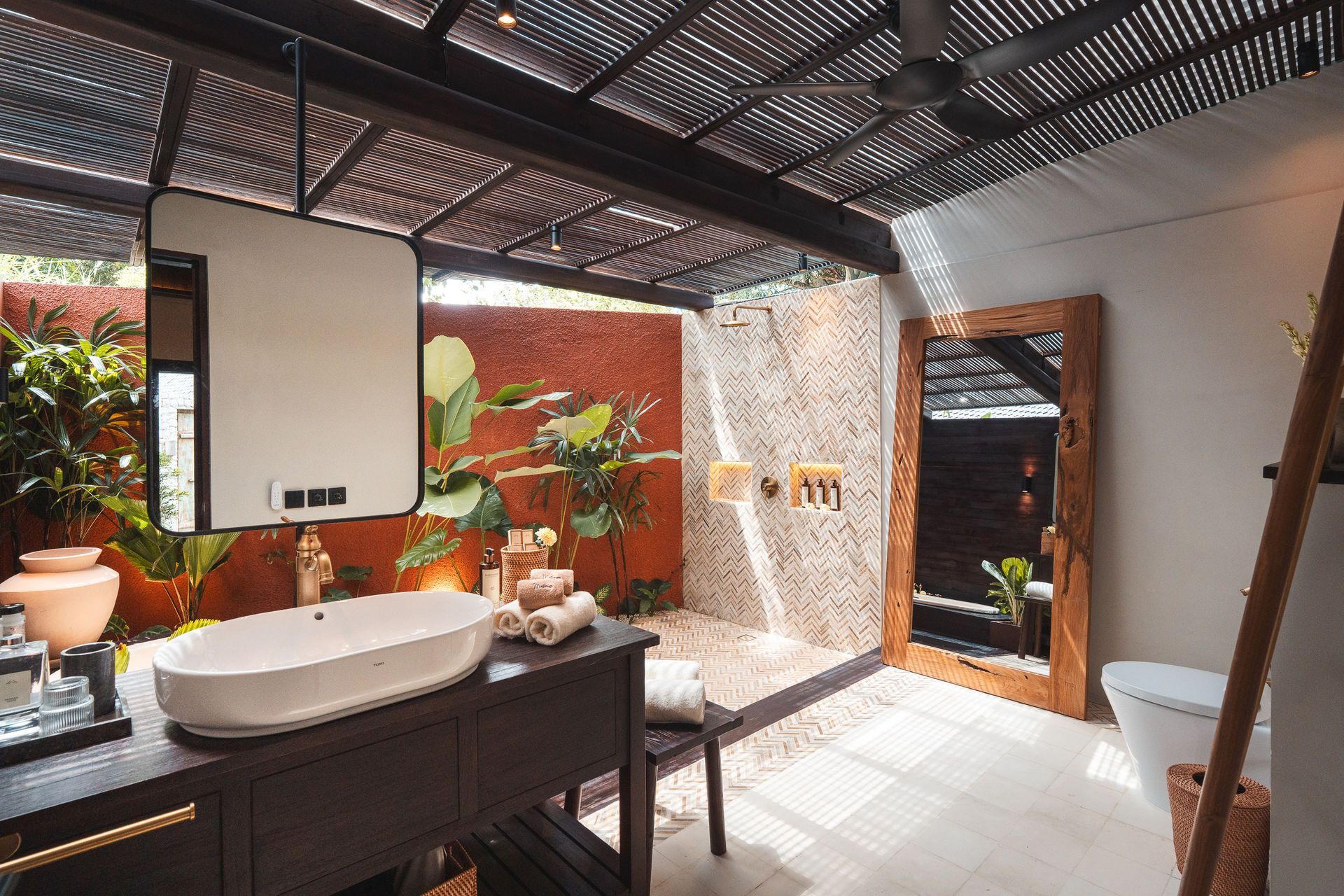 Outdoor bathroom with freestanding tub, vanity, and plants under a slatted roof in bright sunlight