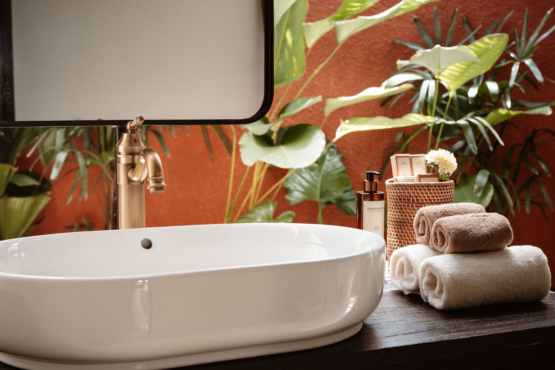 White bathroom sink with gold faucet, round mirror, tropical wallpaper, and stacked towels on the counter