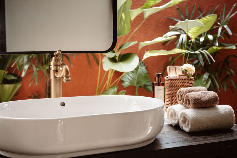 White bathroom sink with gold faucet, round mirror, tropical wallpaper, and stacked towels on the counter