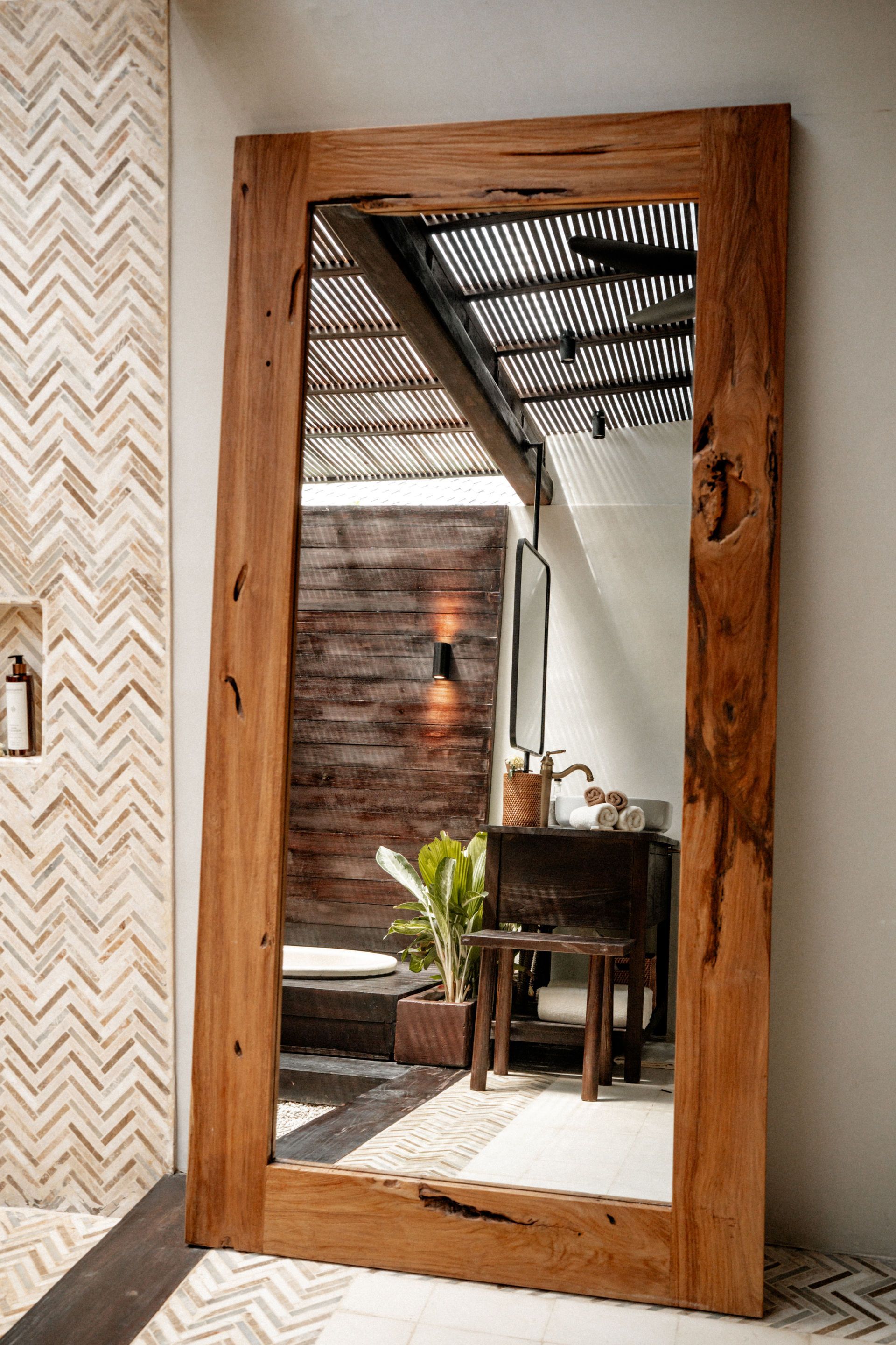 Rustic wooden floor mirror leaning against a wall, reflecting a cozy room with a chair, plant, and wall lights.