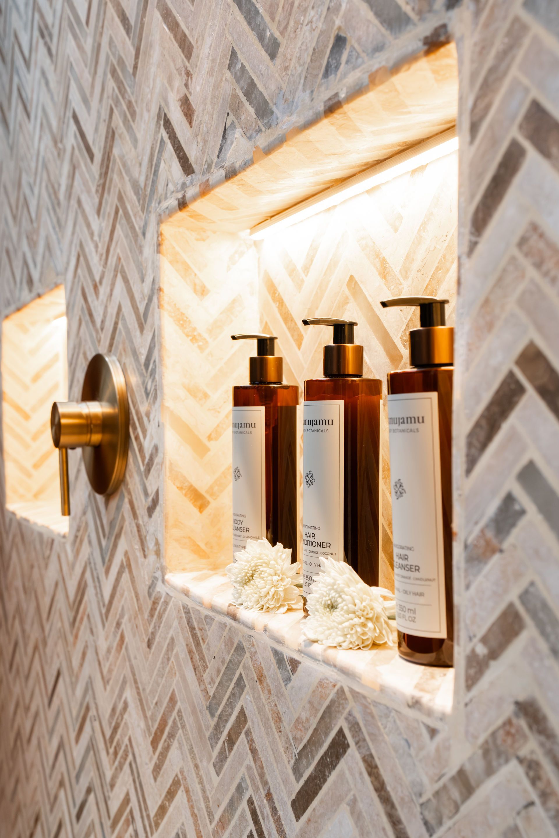Backlit bathroom niche with three amber pump bottles and folded white towels in a patterned tiled wall