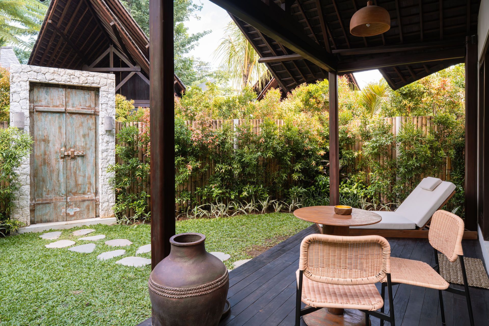Tropical patio with lounge chair, woven chairs, clay jar, and rustic stone door by lush greenery