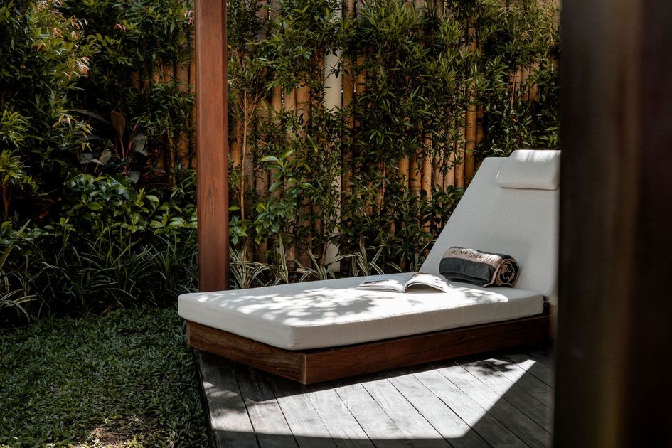 White outdoor lounge chair with a cushion and sunglasses on a wooden deck beside greenery and a fence