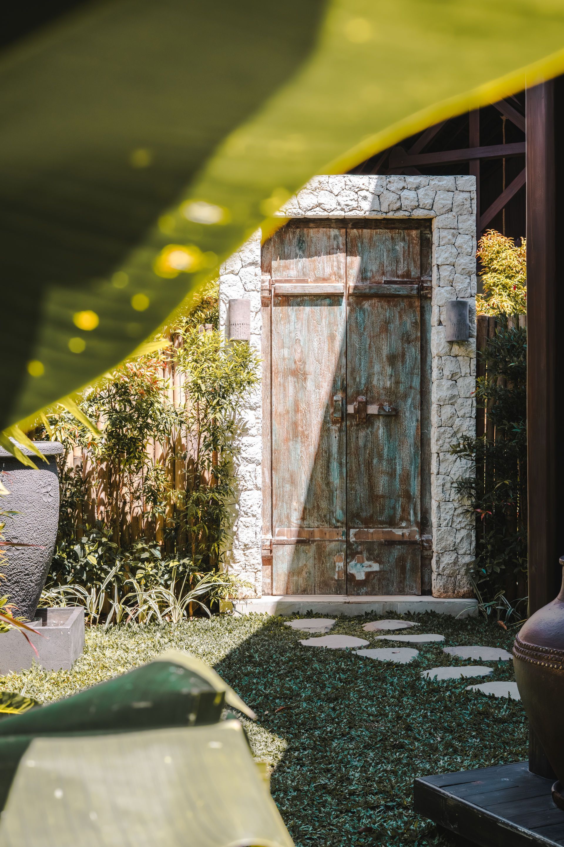 Rustic wooden door set in a stone wall, partially shaded by a green tarp in a sunlit garden.