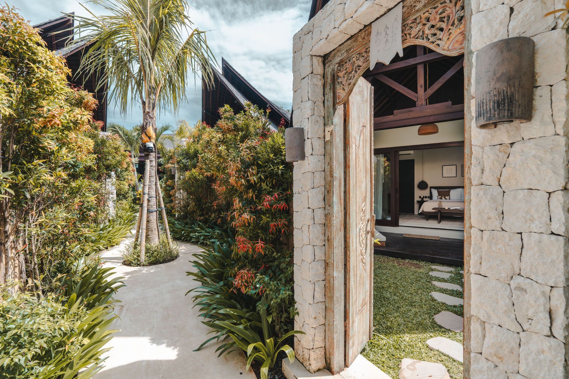 Tropical garden walkway beside a stone building with an open doorway and palm trees.