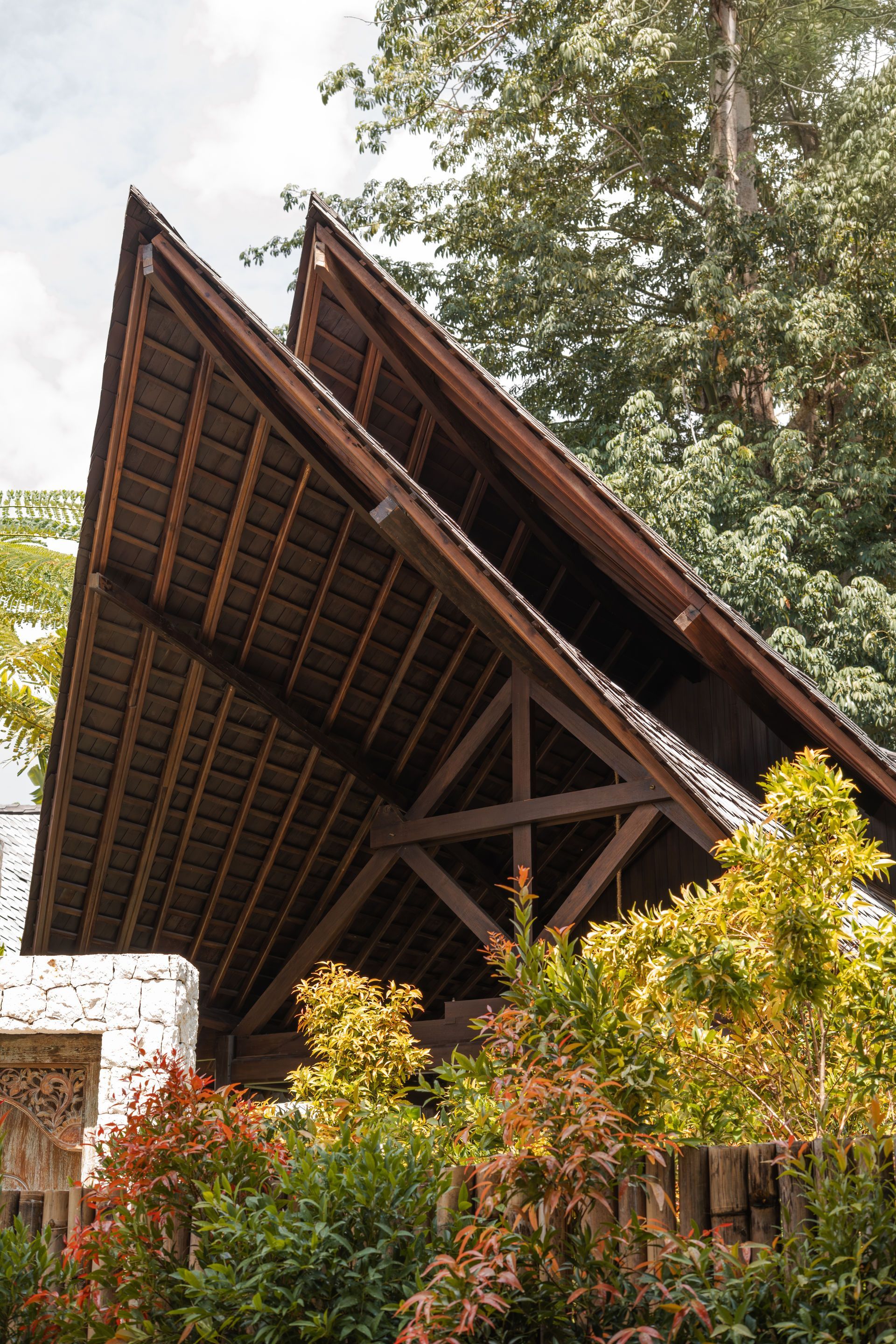 A steeply pitched dark wooden roof above a garden, framed by trees and shrubs.