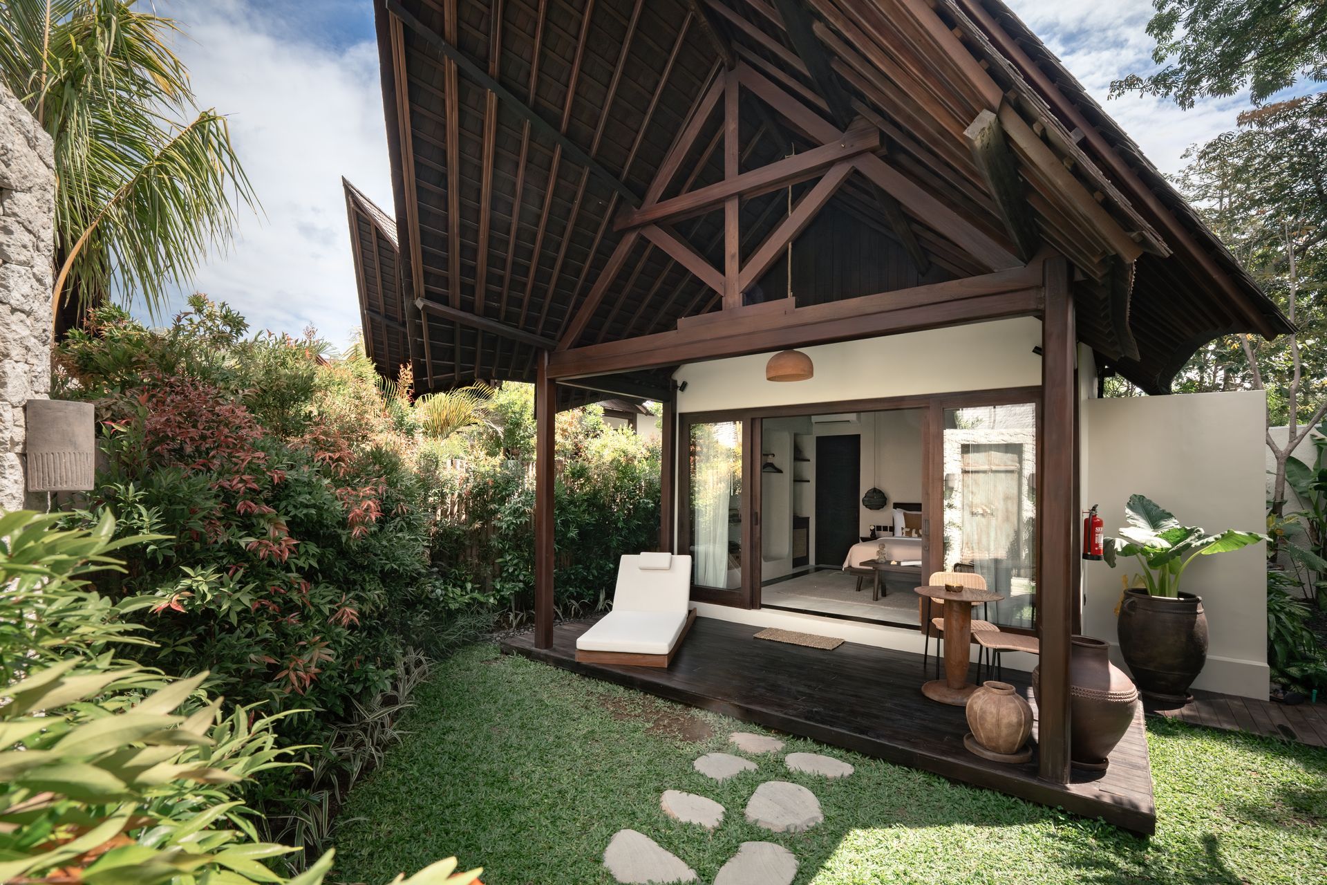 Tropical bungalow porch with thatched roof, white lounge chair, and stone path amid lush greenery