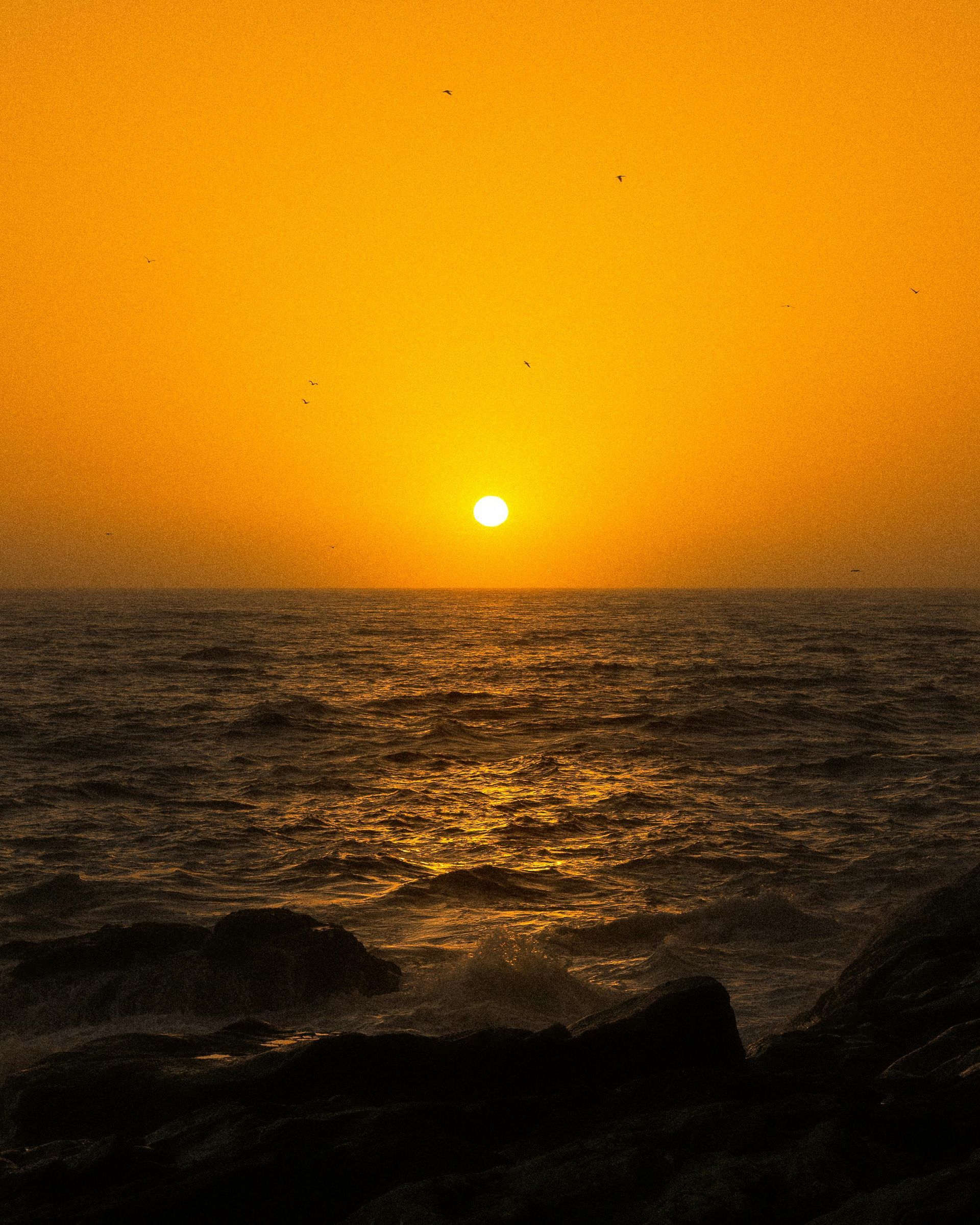 Golden sunset over a dark, rippling ocean with silhouetted rocks in the foreground.