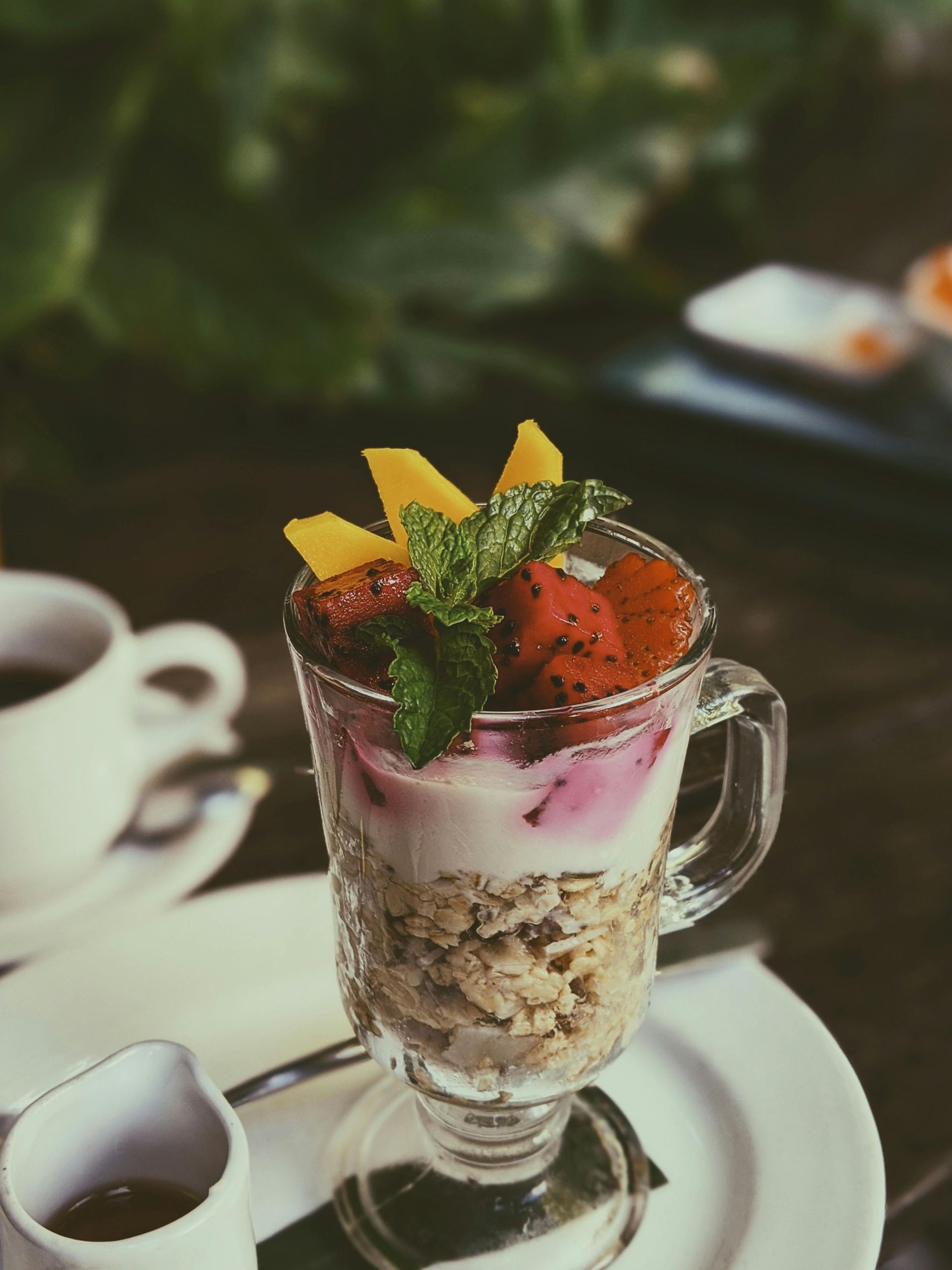 Dessert parfait in a glass mug with strawberries, mint, and banana slices on a saucer beside a coffee cup