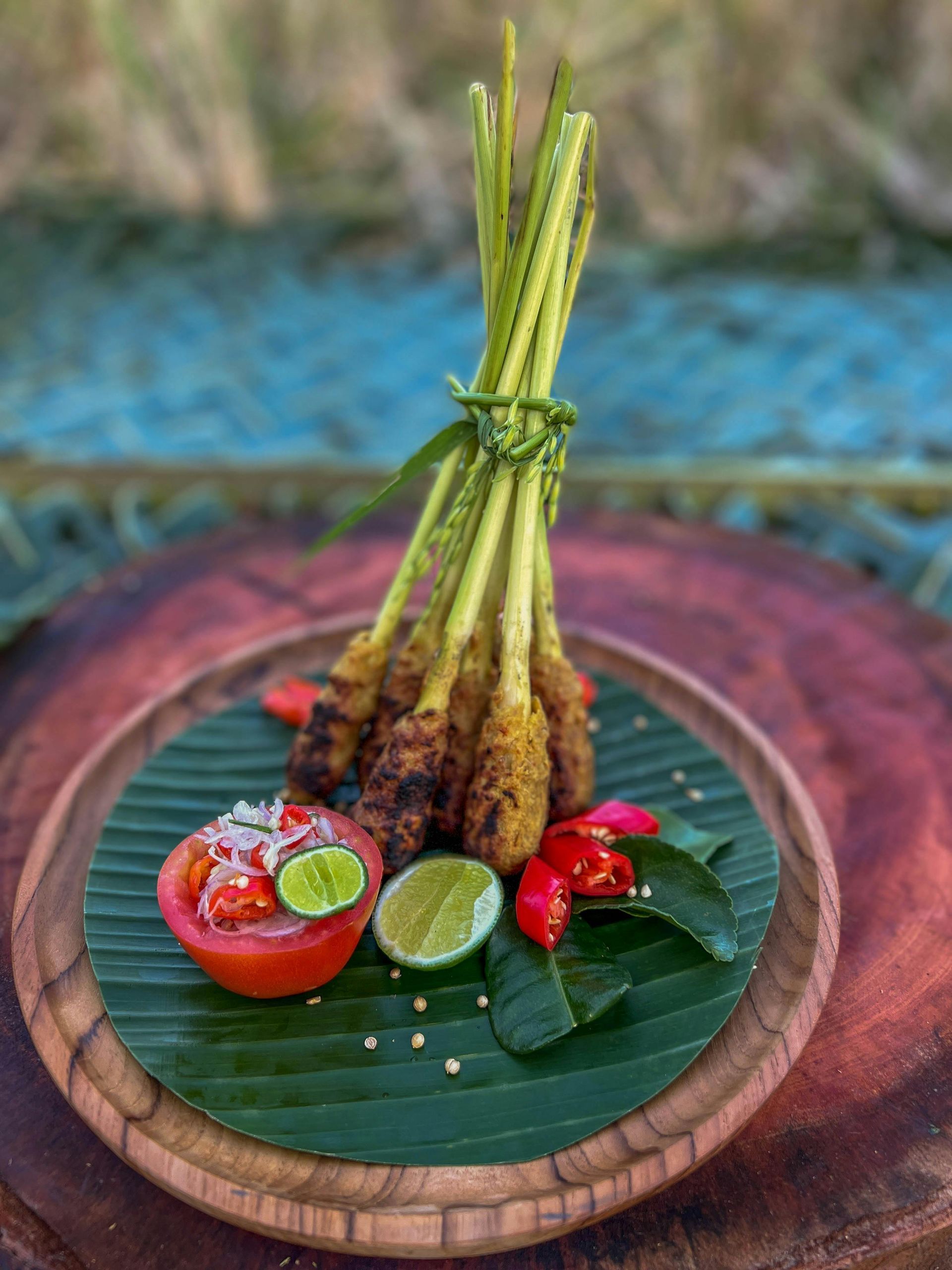 Grilled lemongrass skewers on a banana leaf-lined plate with lime, chili, and tomato garnish