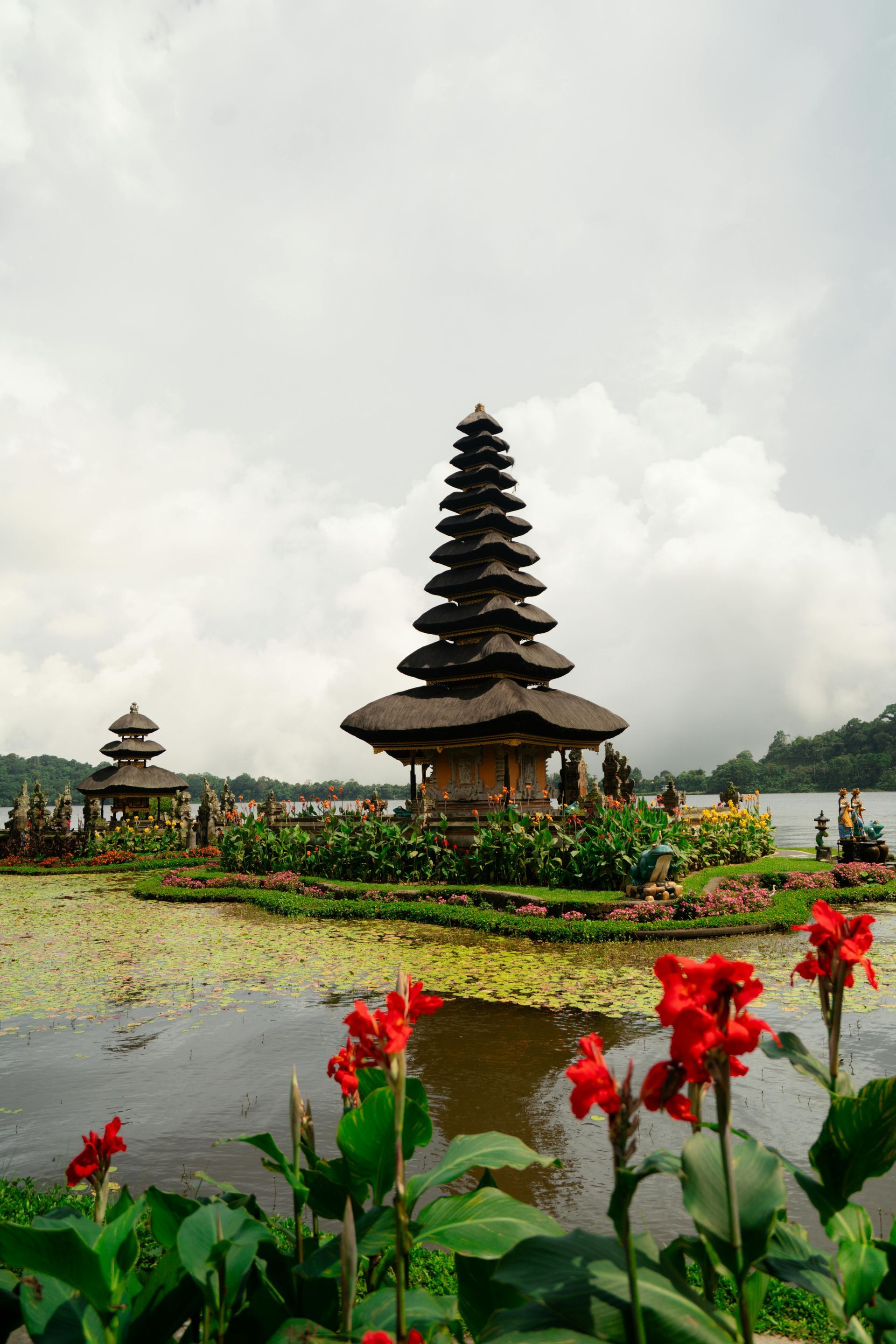 Tall Balinese temple pavilion by a lotus pond, with red flowers in the foreground and a cloudy sky
