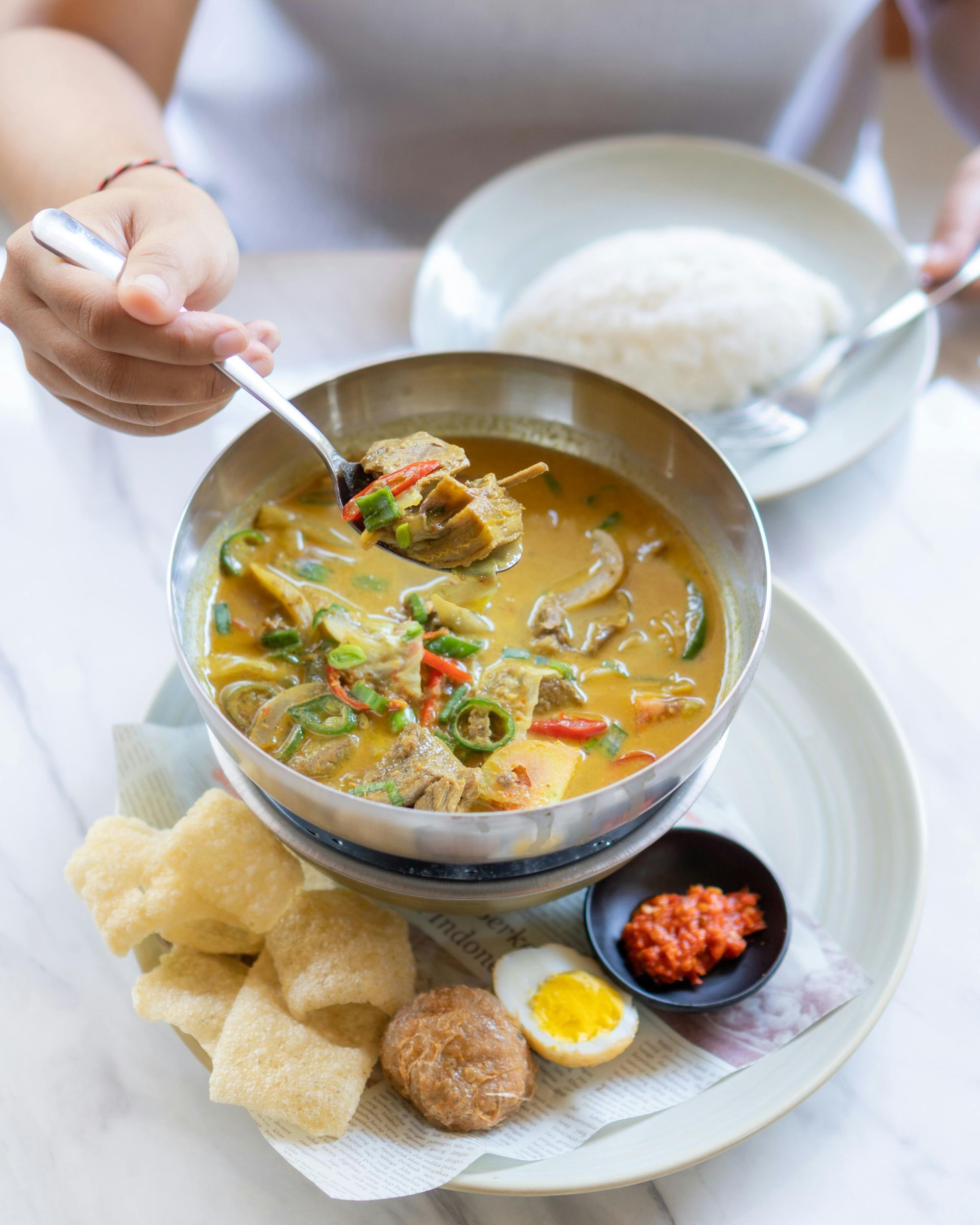 Person serving spicy soup with rice, crackers, and condiments on a white table