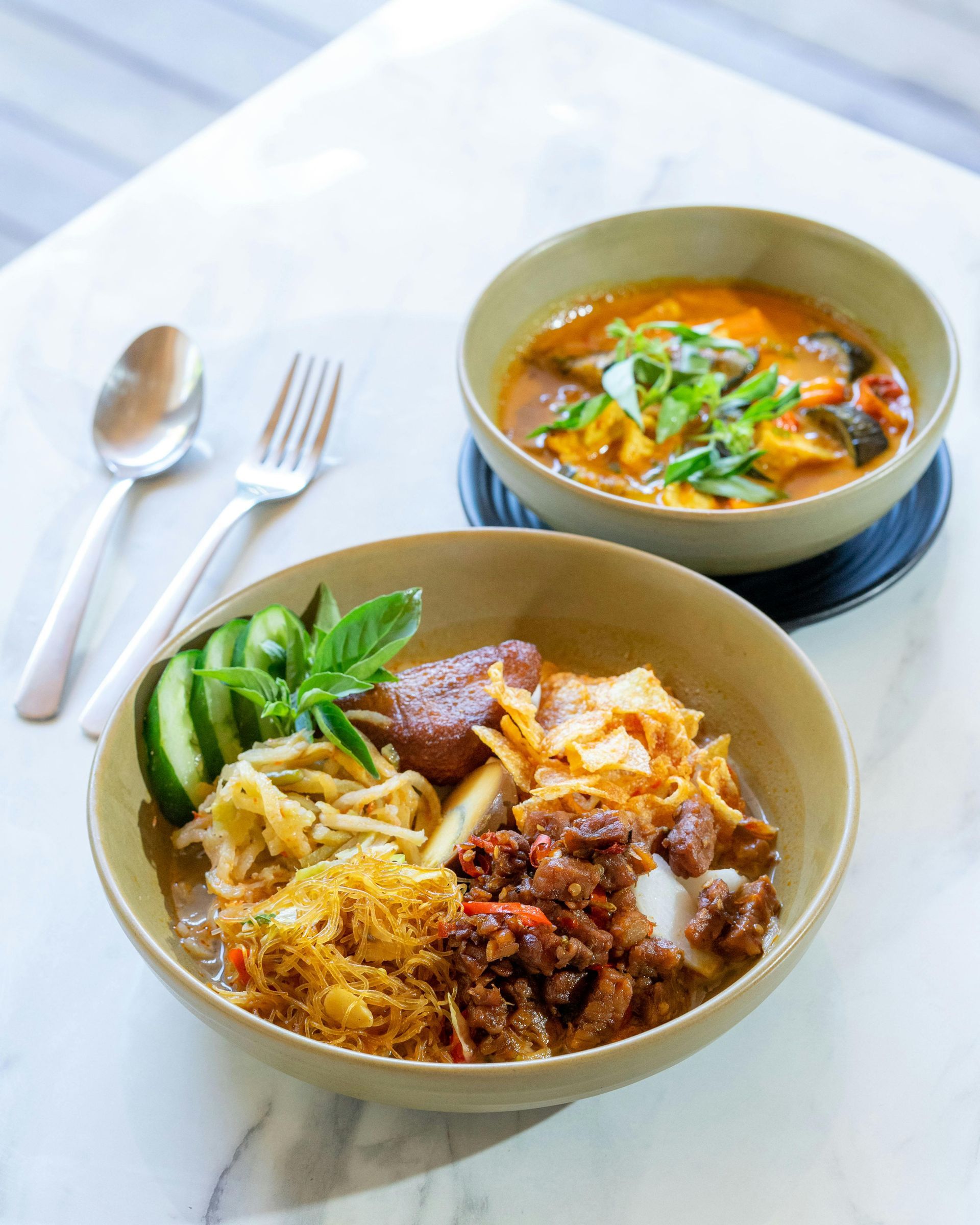 Two bowls of Asian noodle soup and rice dishes with utensils on a light table