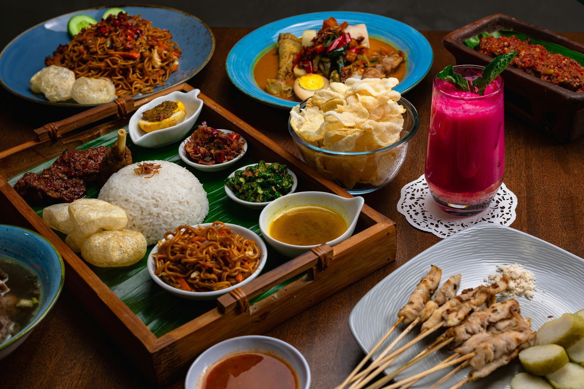 Assorted Indonesian dishes on colorful plates, including rice, skewers, sauces, crackers, and a pink drink on a wooden table