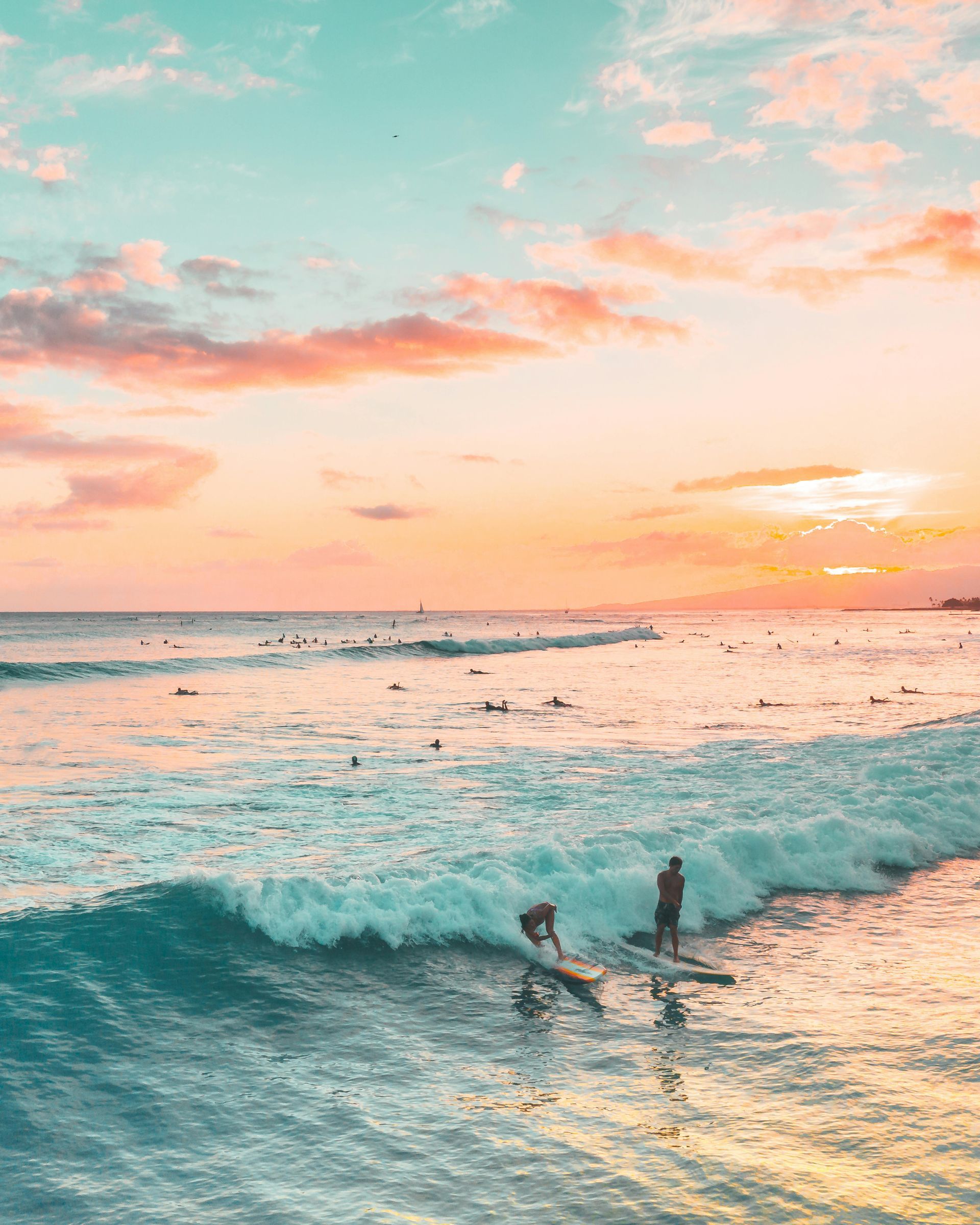 Surfers riding a wave at sunset with pink-orange sky and turquoise water