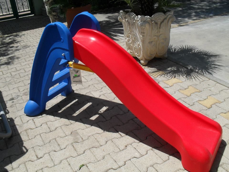 Scivolo per bambini in plastica blu e rossa su un patio in mattoni, vicino a un vaso decorativo.