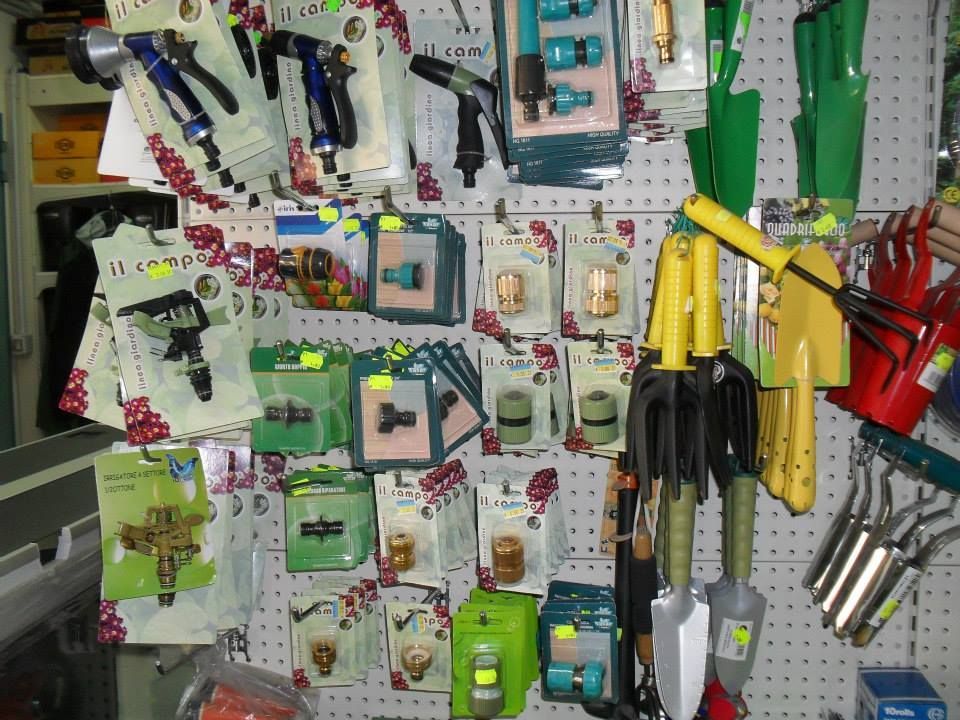 Attrezzi e accessori da giardino esposti su un pannello forato, tra cui spruzzatori, raccordi, cazzuole e rastrelli manuali.