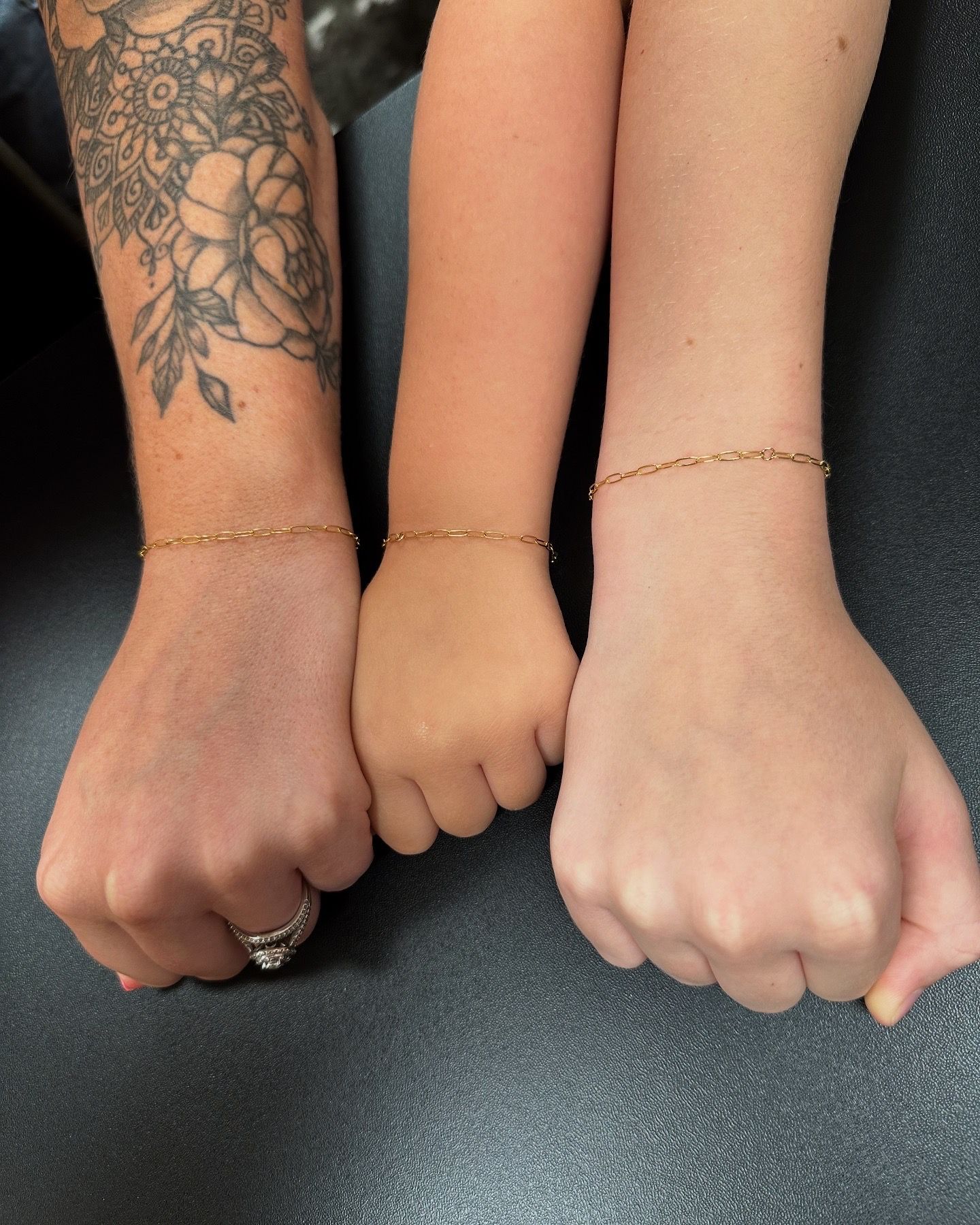 Three wrists wearing gold bracelets are clenched into fists. One arm is tattooed with flowers.