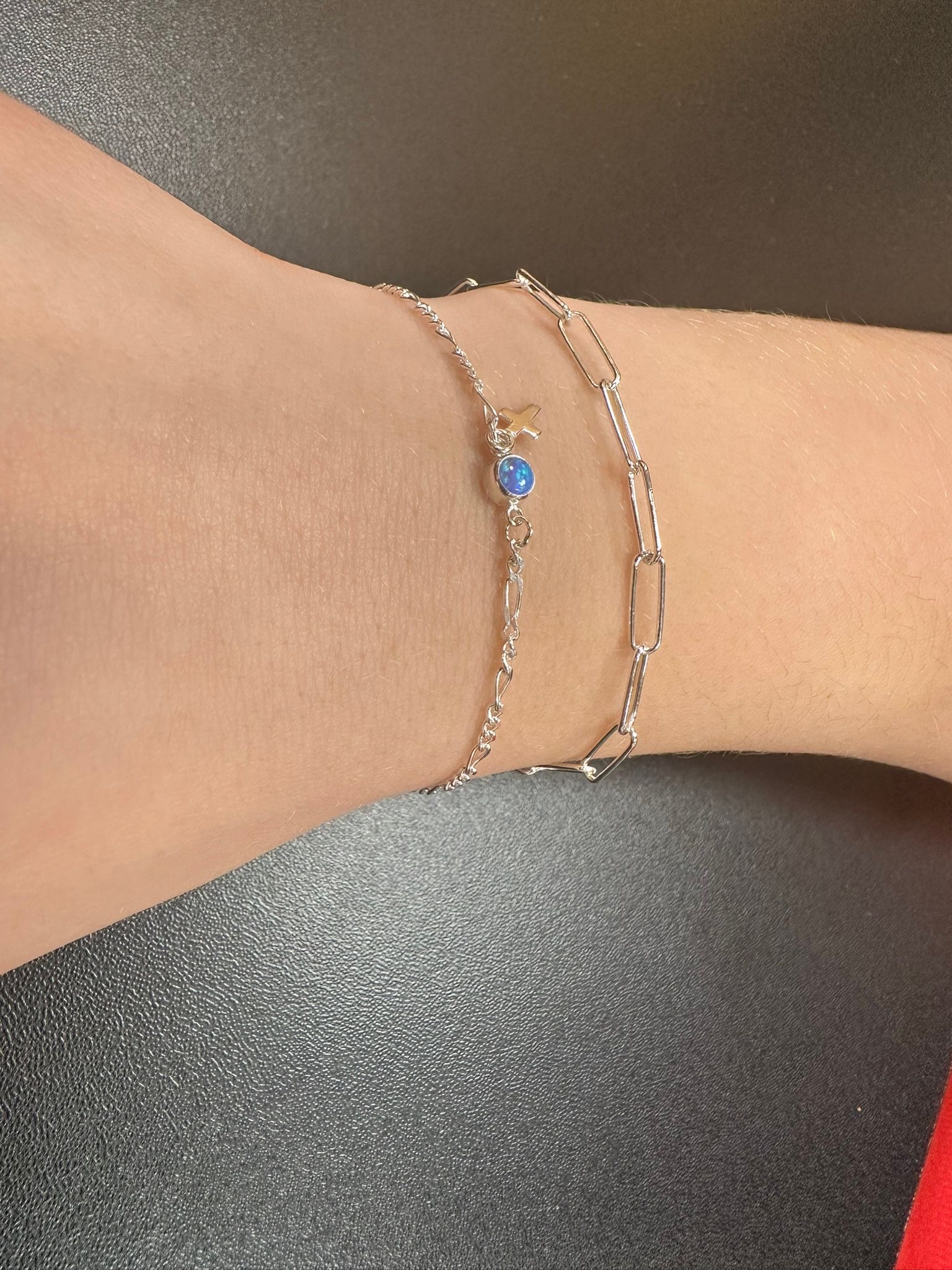 A wrist wearing two delicate silver bracelets; one has a blue gemstone, the other a paperclip chain.
