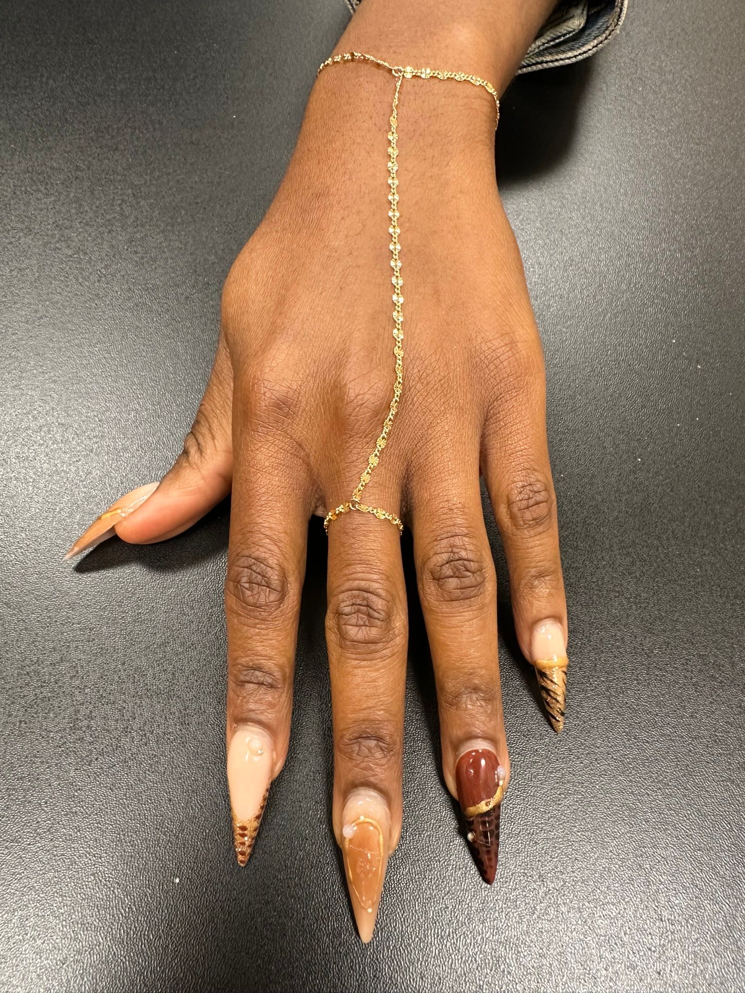 Hand with gold jewelry, long stiletto nails with brown ombre and brown patterned accent.