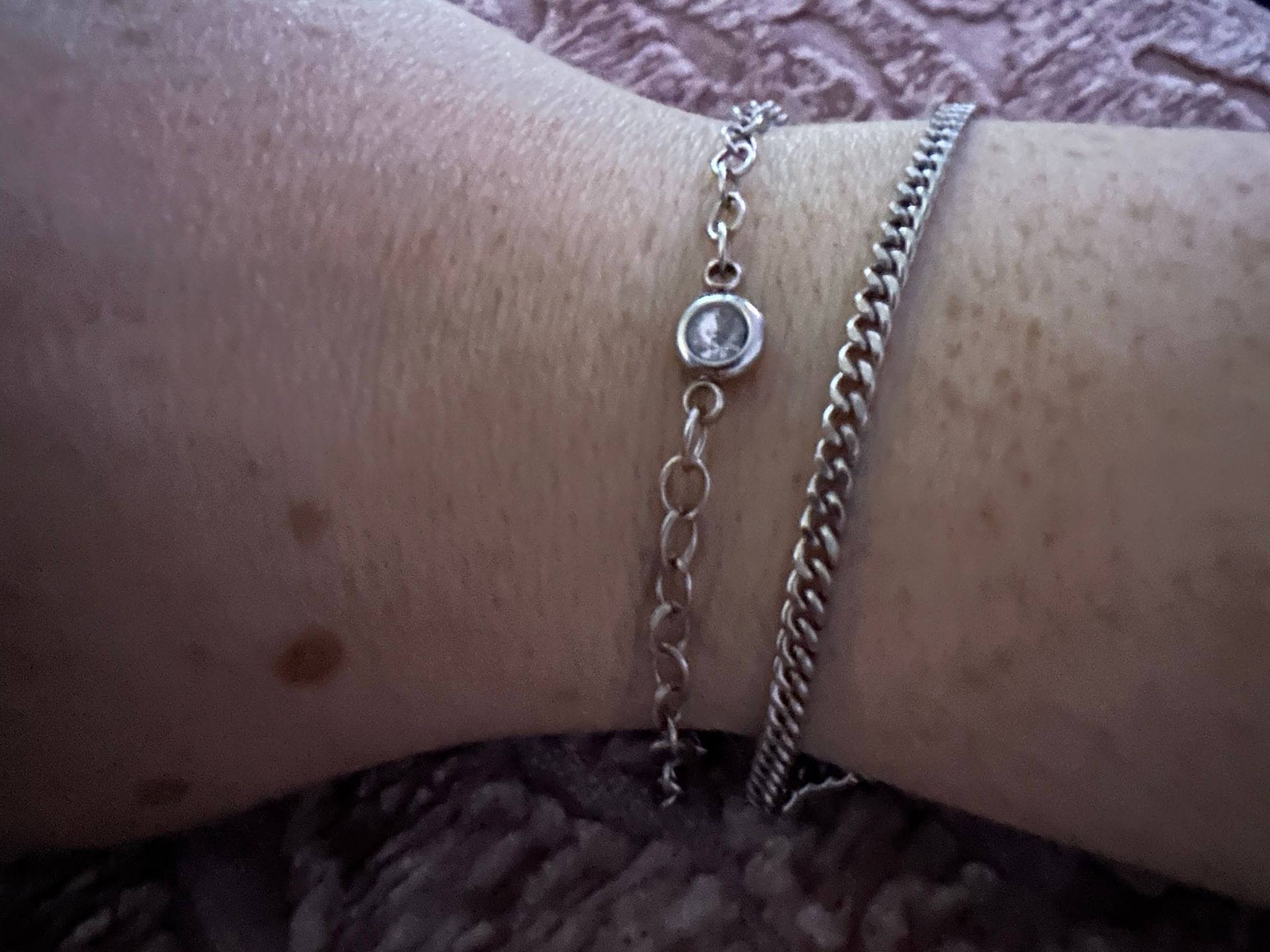 Two silver bracelets on a light-skinned wrist, one with a clear stone, the other a curb chain.
