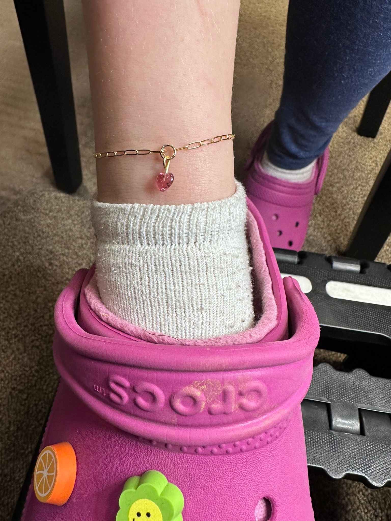 Person's ankle with a gold anklet, gray sock, and pink Crocs. Orange and green shoe charms are visible.